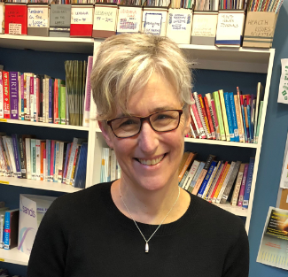 A headshot of a woman with cropped blonde hair, wearing dark glasses, a silver necklace and a black top smiling at the camera. Behind are bookshelves filled with books.