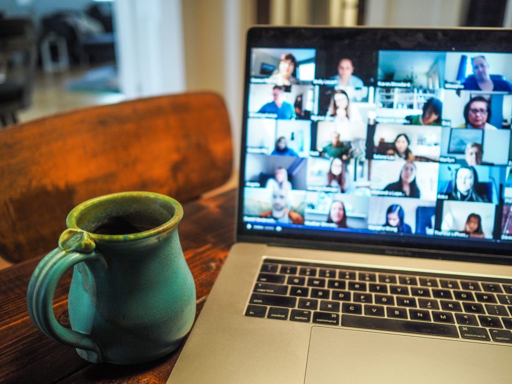 A cropped image of a laptop with faces on the screen attending an online meeting. To the left is a green mug on a brown wooden table with a matching wooden chair.