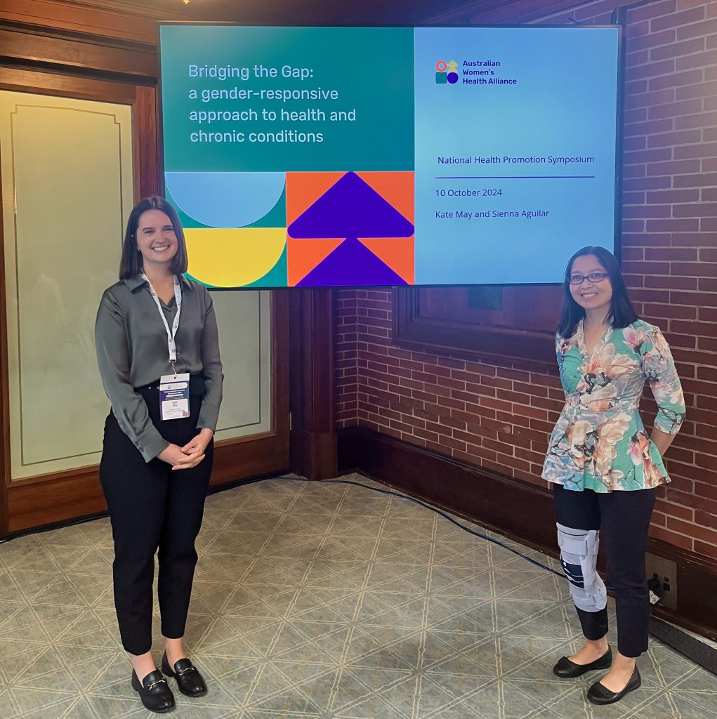 Kate May and Sienna Aguilar from the Australian Women's Health Alliance are standing in front of a digital screen that says: Bridging the Gap: a gender-responsive approach to health and chronic conditions.