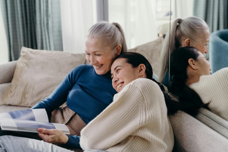 An older white woman with white hair and an Asian woman are smiling and lounging together on a couch.