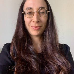 A woman with long brown hair, wearing round glasses and a black top smiles faintly at the camera