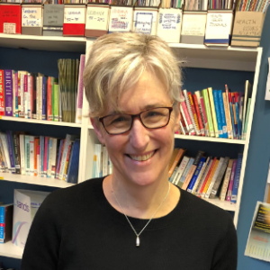 A headshot of a woman with cropped blonde hair, wearing dark glasses, a silver necklace and a black top smiling at the camera. Behind are bookshelves filled with books.