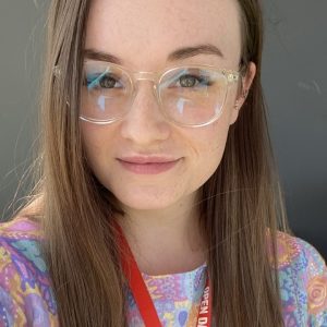 A woman with long light brown hair, wearing clear framed glasses, a silver necklace and a multi-coloured top smiling faintly at the camera.